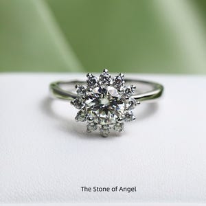 Peut inclure: Une bague en argent ornée d'une grosse pierre précieuse ronde et transparente, entourée de petites pierres, créant un motif floral. La bague est posée sur une surface blanche avec un fond vert. Le texte "The Stone of Angel" est en bas.