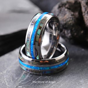 Könnte beinhalten: Zwei silberne Ringe mit einem zentralen Band aus leuchtend blauem Opal und schillernden Abalone-Muscheleinlagen. Die Ringe sind gestapelt, mit dem Text "The Stone of Angel" unten.