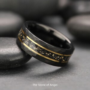 Op de afbeelding: Zwarte ring met een gehamerde textuur, met een goudkleurige inleg met een gespikkeld ontwerp. De ring heeft twee dunne gouden banden. De tekst "The Stone of Angel" staat onderaan de afbeelding.