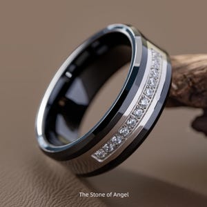 Op de afbeelding: Close-up van een zwart-zilveren ring met een rij kleine, heldere edelstenen. De ring heeft een gepolijste afwerking en de tekst "The Stone of Angel" staat onderaan.