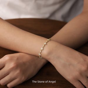 Pode incluir: Uma pulseira dourada com um design floral, adornada com pequenas pedras brilhantes. A pulseira é usada no pulso, com o texto "The Stone of Angel" visível na parte inferior da imagem. É uma peça de joalharia delicada.