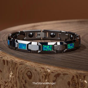 Puede incluir: Una pulsera de eslabones gris oscuro con incrustaciones rectangulares de piedra iridiscente azul y verde vibrante. La pulsera tiene un acabado pulido y un mecanismo de cierre seguro. El texto TheStoneofAngel es visible en la parte inferior.