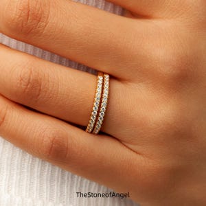 May include: Two thin gold bands featuring a continuous row of small, clear, round-cut stones set along the outer surface. The rings are positioned side-by-side on a finger. The text TheStoneofAngel is printed at the bottom of the image.