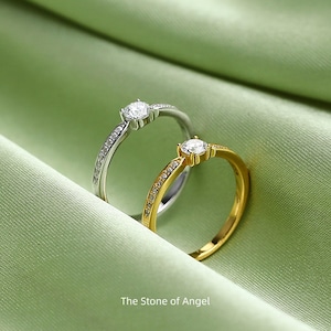 Peut inclure: Deux bagues, une argentée et une dorée, chacune avec une pierre précieuse claire centrale et de petites pierres sur le bandeau. Les bagues reposent sur un tissu de satin vert. Le texte "The Stone of Angel" est en bas.