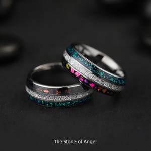 May include: Two silver rings with colorful inlays. Each ring features a band of iridescent green and pink, separated by a textured silver band. The rings are set against a dark background, with the text "The Stone of Angel" at the bottom.