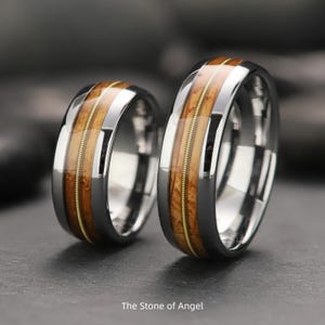 May include: Two matching rings with a polished silver band, a central section of light brown wood, and a thin gold-colored wire inlay. The rings are displayed on a dark surface. The text "The Stone of Angel" is at the bottom.