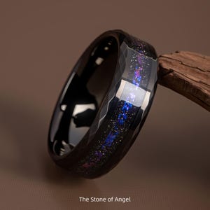 Puede incluir: Anillo negro facetado con una incrustación inspirada en la galaxia con motas azules, moradas y blancas. El anillo tiene una superficie pulida y reflectante. El texto "The Stone of Angel" es visible en la parte inferior.
