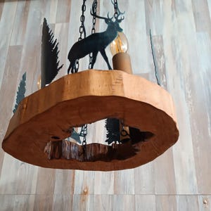 Può includere: Un lampadario rustico in legno con finitura naturale. L'apparecchio presenta una base circolare in legno con un'apertura, una silhouette di cervo in metallo nero e ritagli decorativi di alberi. La lampadina è visibile, sospesa da catene.