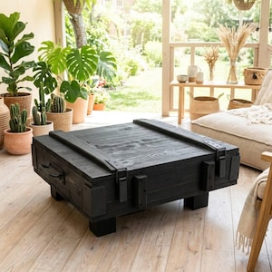 Pode incluir: Uma mesa de centro quadrada construída em madeira tingida de escuro, apresentando um tampo plano com duas ripas de madeira paralelas, suportes de canto em metal preto e pegas laterais. A mesa assenta em quatro pés de bloco robustos.