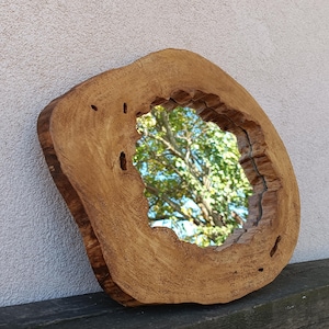 Op de afbeelding: Een ronde houten spiegel met een natuurlijke houten afwerking. De spiegel is in een cirkelvormig stuk hout gezet met een ruwe, organische rand. De spiegel reflecteert een uitzicht op bomen en bladeren.