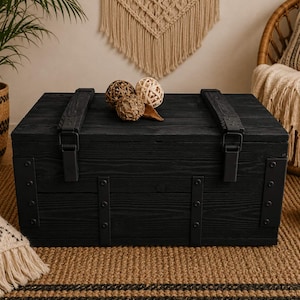 May include: A rectangular black wooden storage chest featuring a hinged lid with two heavy-duty metal latches. The front and top panels are reinforced with vertical black metal straps secured by visible rivets, showcasing a prominent wood grain texture.