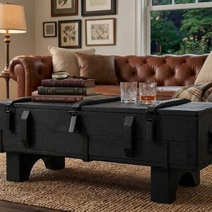Può includere: Tavolino nero in legno a forma di baule con ferramenta in metallo. Il tavolo ha una forma rettangolare e poggia su quattro gambe corte. Il tavolo si trova in un soggiorno con un divano in pelle marrone e un tappeto in juta.