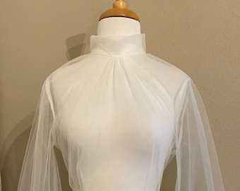 Puede incluir: Top blanco transparente de manga larga y cuello alto. El top tiene un corpiño ajustado y un detalle fruncido en el cuello. Las mangas están fruncidas en los puños.