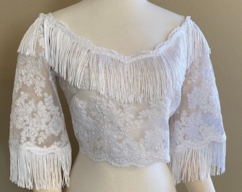 Puede incluir: Top blanco de encaje de hombros descubiertos con ribete de flecos blancos alrededor del escote y las mangas.
