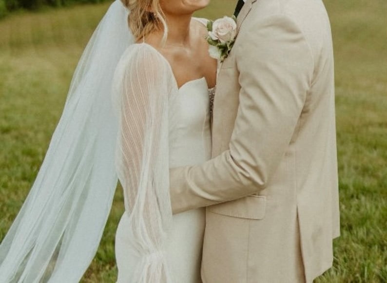 Puede incluir: Una novia con un vestido de novia blanco con mangas transparentes y un velo, abrazando a una persona con un traje color beige. Un ojal de rosa adorna el traje. La pareja est&aacute; en un campo de hierba.