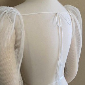 May include: White sheer top with puffed sleeves and a tie at the back. The top has a satin ribbon around the waist.