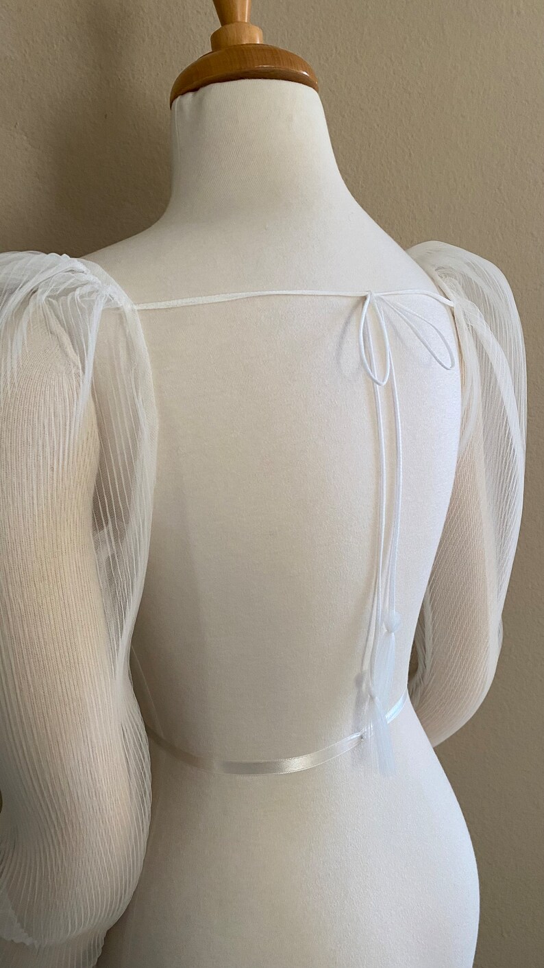 May include: White sheer top with puffed sleeves and a tie at the back. The top has a satin ribbon around the waist.
