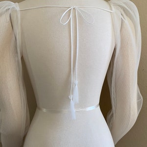 May include: White sheer top with puffed sleeves and a tie at the back. The top has a white ribbon around the waist.