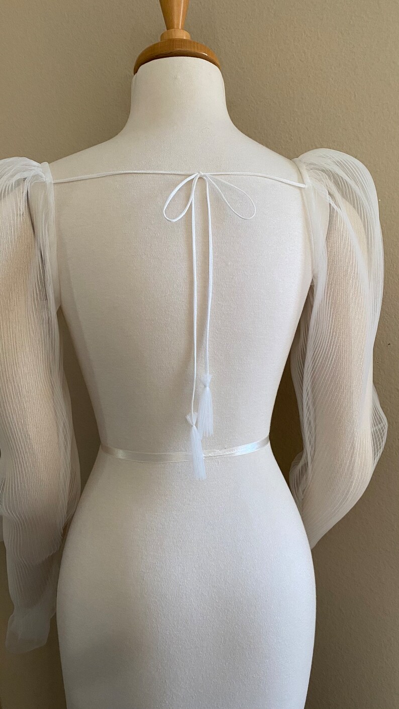 May include: White sheer top with puffed sleeves and a tie at the back. The top has a white ribbon around the waist.