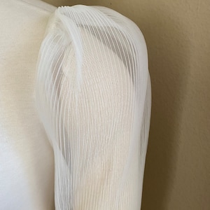 May include: White sheer fabric with a pleated texture. The fabric is gathered at the top and has a loose, flowing shape.