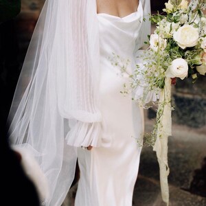 Puede incluir: Vestido de novia blanco con cuello drapeado y mangas transparentes plisadas. La novia sostiene un ramo de flores blancas y crema con cintas. Un velo transparente cubre los hombros.