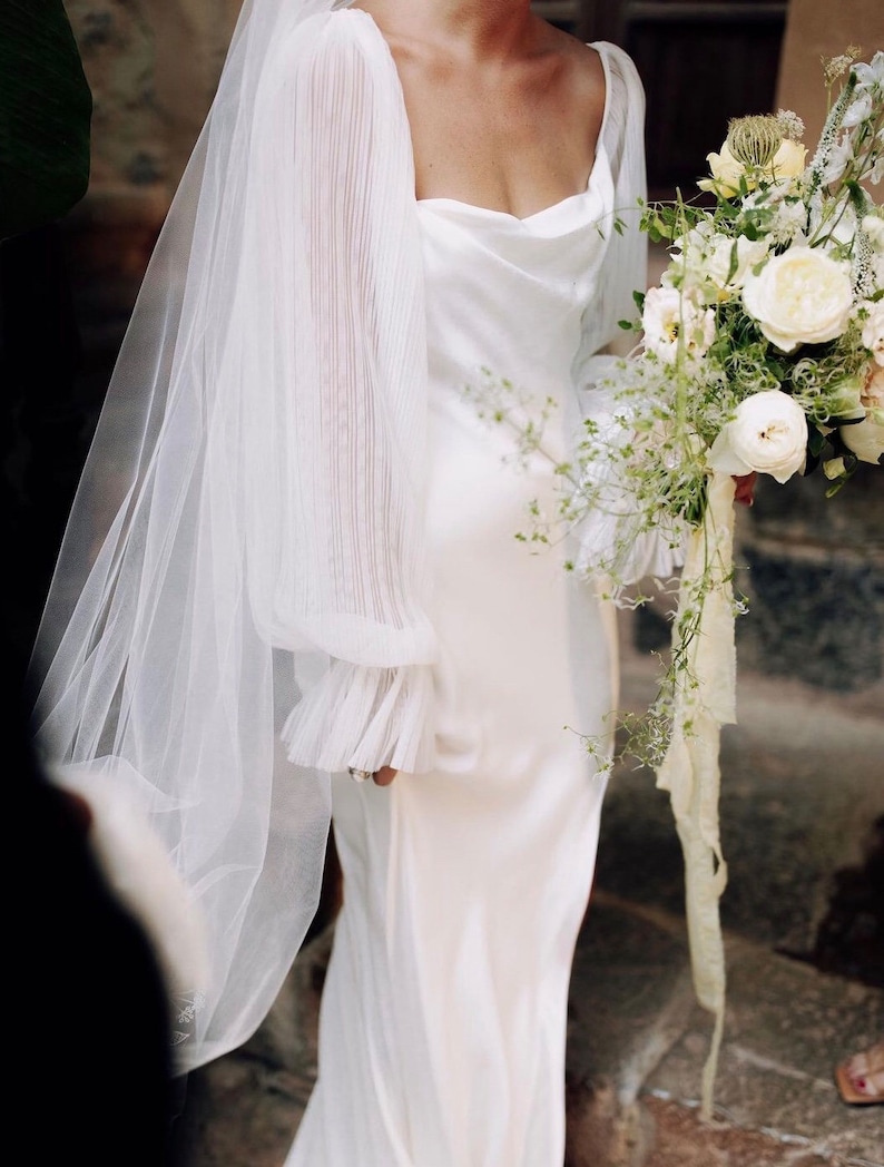 Puede incluir: Vestido de novia blanco con cuello drapeado y mangas transparentes plisadas. La novia sostiene un ramo de flores blancas y crema con cintas. Un velo transparente cubre los hombros.