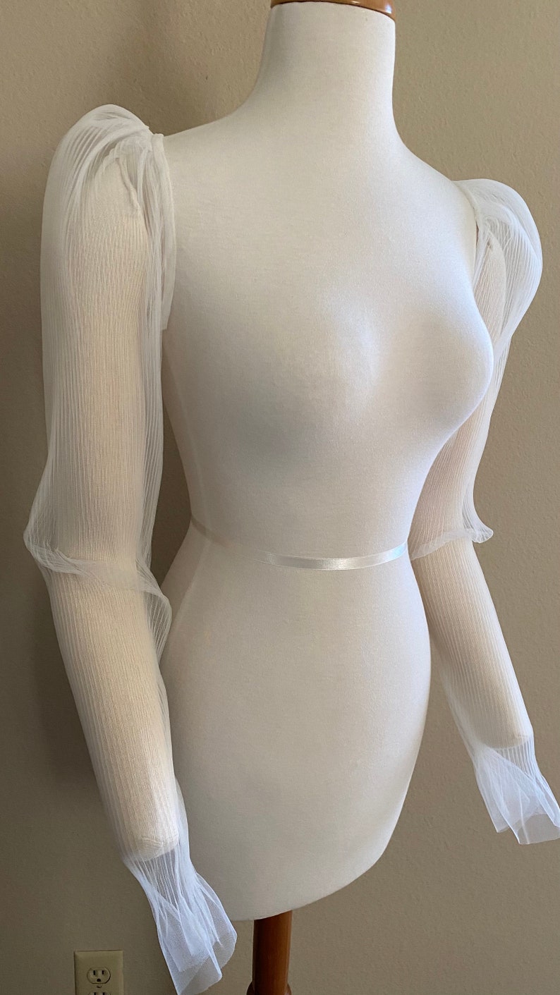May include: White sheer fabric sleeves with a gathered puff at the shoulder and a ruffled cuff. The sleeves are on a white dress form.