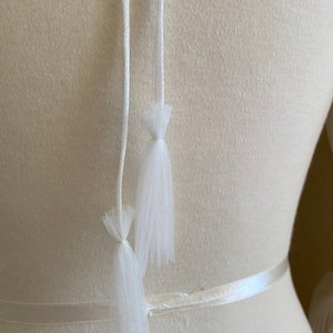 May include: White tulle ribbon ties with a white satin ribbon. The ties are attached to a white dress form.