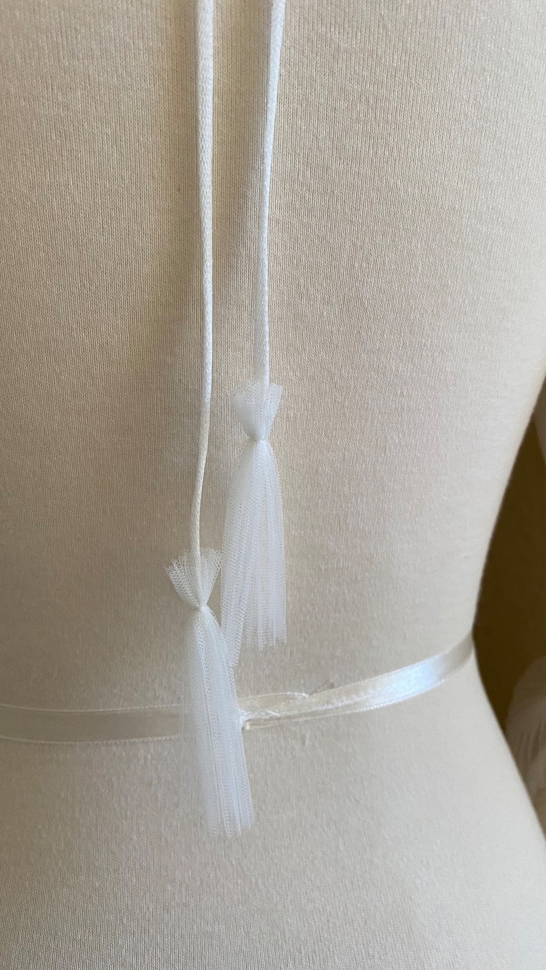 May include: White tulle ribbon ties with a white satin ribbon. The ties are attached to a white dress form.