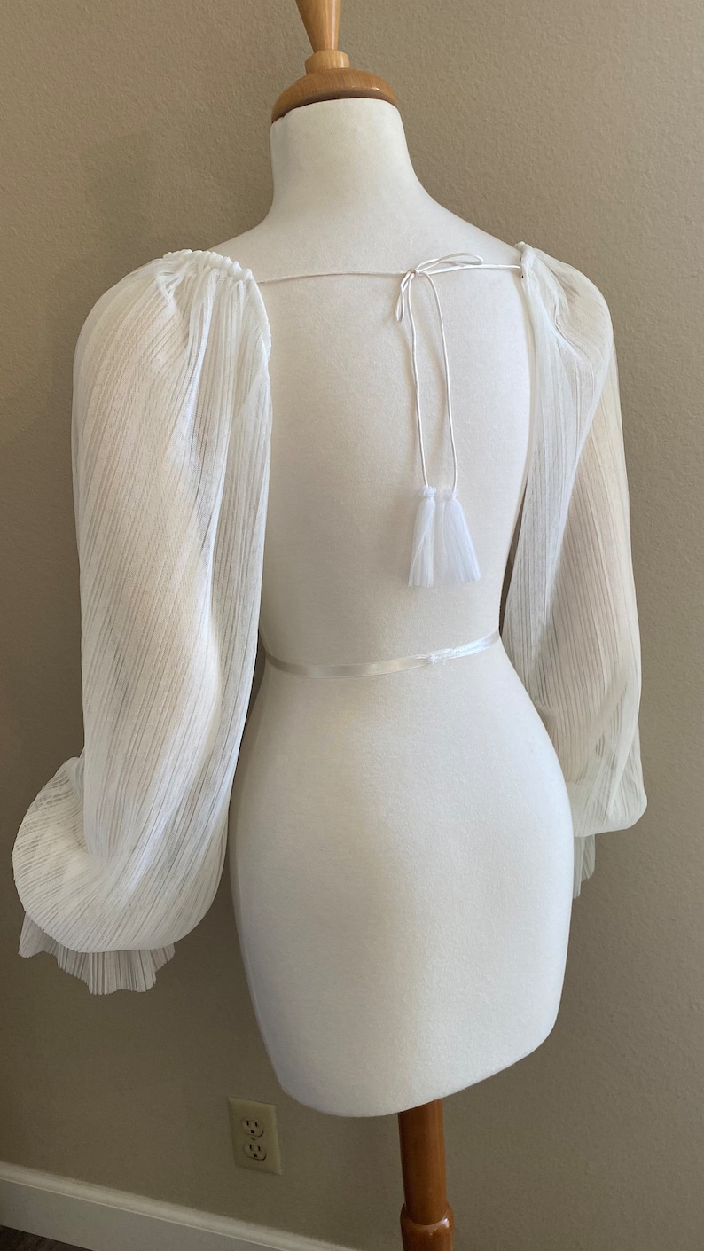 Puede incluir: Top blanco transparente de manga larga, cuello cuadrado y cierre de lazo en la espalda. Las mangas est&aacute;n recogidas en los hombros y las mu&ntilde;ecas, creando una silueta voluminosa y elegante. Detalle de borla blanca en la espalda.