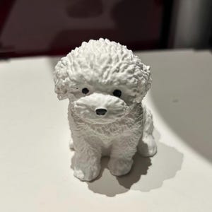 May include: White ceramic figurine of a small dog with fluffy fur. The dog is sitting with its head tilted down and its eyes closed.