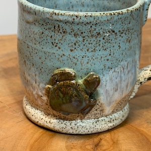 Könnte beinhalten: Ein Keramikbecher mit gesprenkelter hellblauer und weißer Glasur, der auf der Vorderseite ein erhabenes, dreidimensionales dunkelgrünes Schildkrötenemblem aufweist. Der Becher hat einen zylindrischen Körper, einen breiten Rand und eine texturierte Oberfläche.