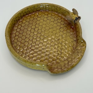 Op de afbeelding: Een keramische lepelhouder met een honingraatpatroon in geel- en bruintinten. Een klein bijenfiguur is aan de rand bevestigd. De lepelhouder is rond met een kleine inkeping voor de lepel.
