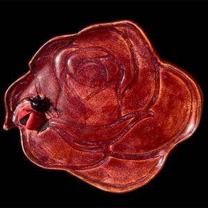 May include: A ceramic dish in the shape of a rose, glazed in a deep reddish-brown color. A small ladybug figurine rests on the edge of the rose. The dish has a glossy finish and is set against a black background.
