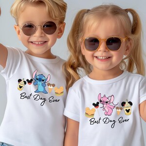 Two young children wearing white t-shirts with the text "Best Day Ever" and cartoon characters, smiling and making a peace sign gesture.