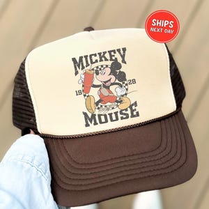 May include: A brown and tan trucker hat featuring a Mickey Mouse graphic. The graphic includes the text "Mickey Mouse" and the year "1928". The hat has a brown brim and mesh backing.