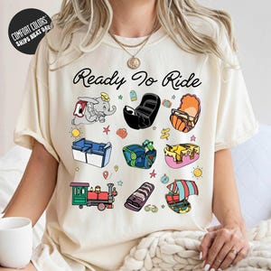 A woman wearing a white t-shirt with a graphic design featuring various cartoon-style illustrations of different objects and activities, along with the text "Ready to Ride".
