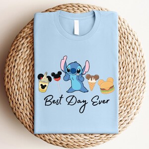A light blue t-shirt with a cartoon character and various Disney-themed items, such as Mickey Mouse and a hamburger, along with the text "Best Day Ever".