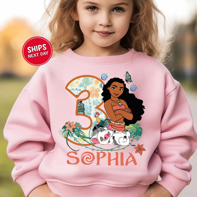 A young girl wearing a pink sweatshirt with a cartoon image of a girl and a number 3 on it.