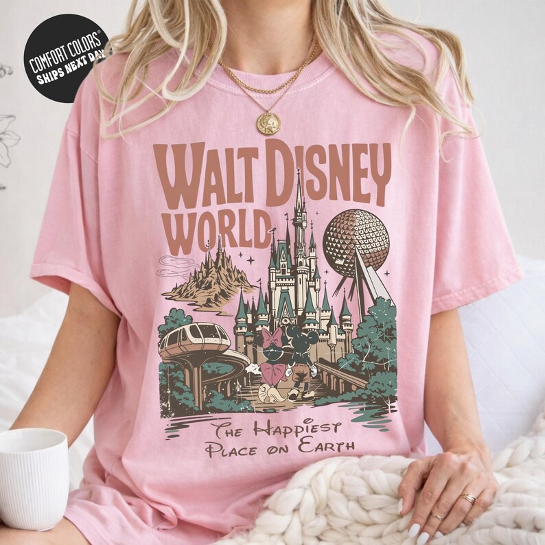 A woman wearing a pink t-shirt with a graphic design of Disney World, including a castle, roller coaster, and other iconic landmarks.