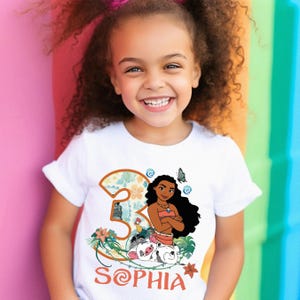 A young girl with curly hair is smiling and wearing a white t-shirt with a graphic of a character from the movie Moana.