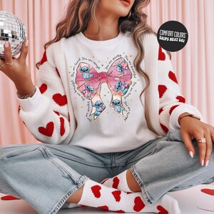 A woman sitting on the floor wearing a white sweater with a bow and red hearts, holding a disco ball in one hand and a heart-shaped object in the other.