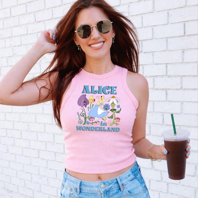 A woman wearing a pink tank top with a graphic of Alice in Wonderland and sunglasses is holding a drink in her hand.