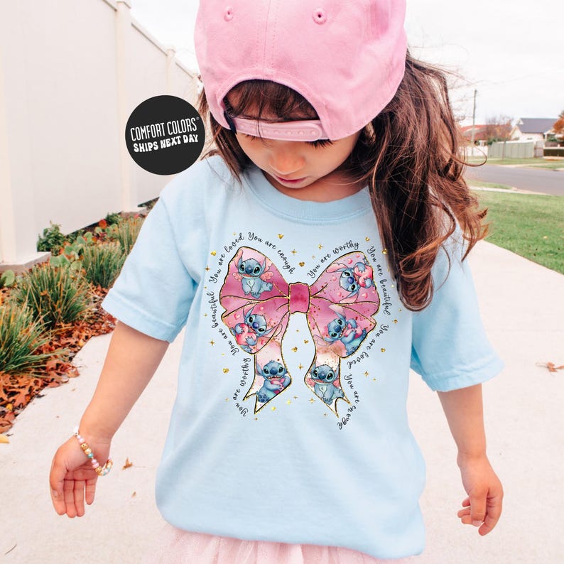 A young girl wearing a pink hat and a light blue t-shirt with a large bow and various cartoon characters on it, standing on a sidewalk.