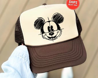 May include: A brown and tan trucker hat featuring a sketched Mickey Mouse design. The hat has a brown brim and mesh back, with a cream-colored front panel displaying the black and white cartoon character. A red circle with the text "SHIPS NEXT DAY" is visible.