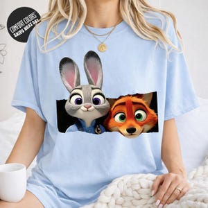 A woman wearing a light blue t-shirt with a graphic of a rabbit and a fox peeking out from behind a black hole.