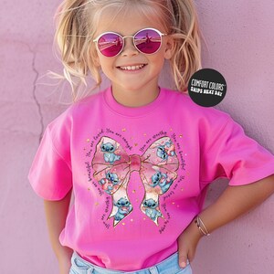 A young girl wearing a pink t-shirt with a bow and sunglasses, smiling at the camera.