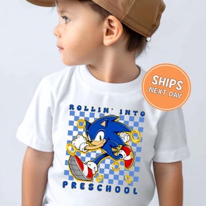 May include: White t-shirt featuring a blue cartoon character with gold rings, set against a blue and white checkered background. The text "ROLLIN' INTO PRESCHOOL" is printed above and below the character.