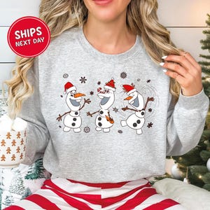 May include: A light gray sweatshirt featuring a festive design with three cartoon snowmen wearing Santa hats and surrounded by snowflakes. The snowmen have carrot noses and stick arms. The sweatshirt has a crew neck.