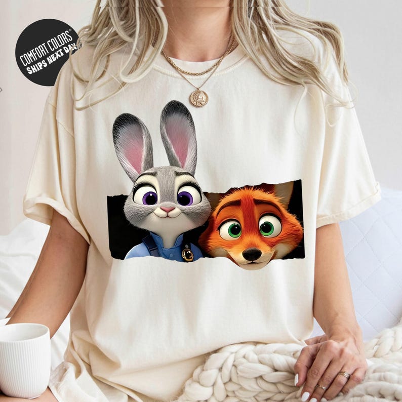 A person wearing a white t-shirt with a graphic of two cartoon animal characters, a rabbit and a fox, peeking out from behind a black background.